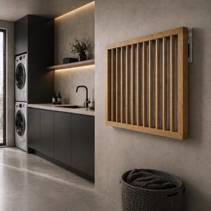 Könnte beinhalten: Ein Wäschetrockner aus Holz mit vertikalen Lamellen ist an einer grauen Wand in einem modernen Waschraum montiert. Der Trockner ist rechteckig und aus hellem Holz gefertigt. Ein dunkelgrauer Wäschekorb steht darunter.
