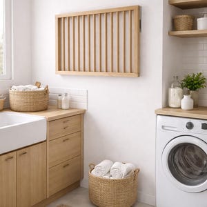 Puede incluir: Un tendedero de madera clara está montado en una pared blanca en una zona de lavandería. Cestas de mimbre contienen toallas dobladas. Una lavadora blanca y armarios de madera completan la habitación.