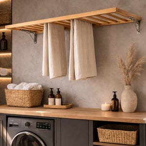 Könnte beinhalten: Ein moderner Waschraum mit einem hölzernen Trockengestell, das zwei beige Handtücher hält. Unter einer Holz-Arbeitsplatte befindet sich eine Waschmaschine. Weidenkörbe, eine Vase und Flaschen ergänzen die Einrichtung. Der Raum hat eine neutrale Farbpalette.