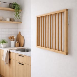 Puede incluir: Un escurreplatos rectangular de madera clara con listones verticales, montado en una pared blanca. El escurreplatos está diseñado para secar la vajilla. El entorno de la cocina incluye estantes, un fregadero y armarios de madera.