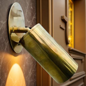 Può includere: Un faretto da parete color ottone. Il faretto ha una base circolare e un alloggiamento cilindrico. La luce è angolata verso il basso, proiettando un bagliore caldo. Il faretto è montato su una parete con un motivo marmorizzato.