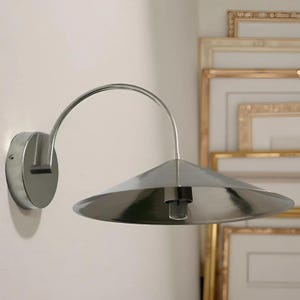Op de afbeelding: Zilverkleurige wandlamp met een gebogen arm en een conische kap. De metalen armatuur is bevestigd aan een witte muur. Modern en minimalistisch ontwerp, geschikt voor diverse interieurstijlen.