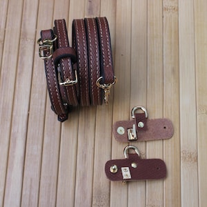 May include: Brown leather bag straps with gold-colored hardware, including buckles and clasps. The straps are stitched with white thread. Two additional leather pieces with clasps are also visible.