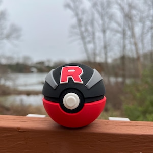 May include: A red, black, and gray novelty ball with a white button and the letter "R" in red. The ball is resting on a brown wooden surface with a blurred outdoor background.