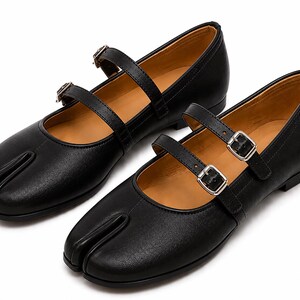 May include: A pair of black leather Tabi shoes with a split toe design. The shoes have two straps with silver buckles and a tan interior. The shoes are flat and have a classic Mary Jane style.