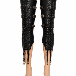 May include: A pair of black thigh-high leg covers featuring vertical lace-up detailing with metal eyelets, three horizontal straps with bronze-toned buckles across the front, and two dangling straps with metal clips at the bottom hem.