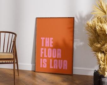 Puede incluir: Un póster naranja enmarcado con el texto "THE FLOOR IS LAVA" en letras rosas. El póster está apoyado contra una pared blanca sobre un suelo de madera. Una silla de madera está a la izquierda del póster.