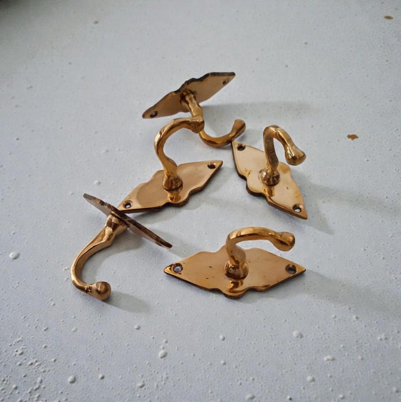 May include: Four antique brass wall hooks. Each hook has a curved design and is mounted on a decorative, diamond-shaped backplate. The hooks are a warm, golden color and appear to be vintage or reclaimed.