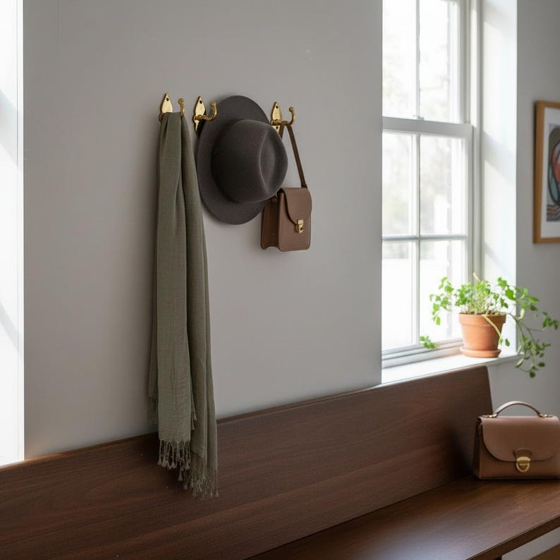 May include: A wall-mounted brass hook rack holds a gray fedora, a small brown purse, and a light green scarf. The hooks are mounted on a light gray wall above a dark brown wooden bench. A brown purse sits on the bench.