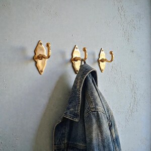 May include: Three brass wall hooks with a decorative design are mounted on a light gray wall. A denim jacket hangs from one of the hooks. The hooks are made of brass and have a curved hook shape.