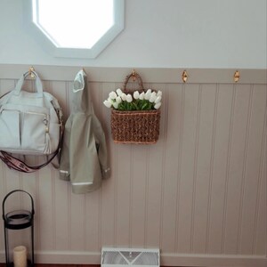 May include: A light gray handbag with a patterned strap hangs on a brass hook next to a light green raincoat. A woven basket filled with white tulips is suspended from a hook. A black lantern with a candle sits on the floor.
