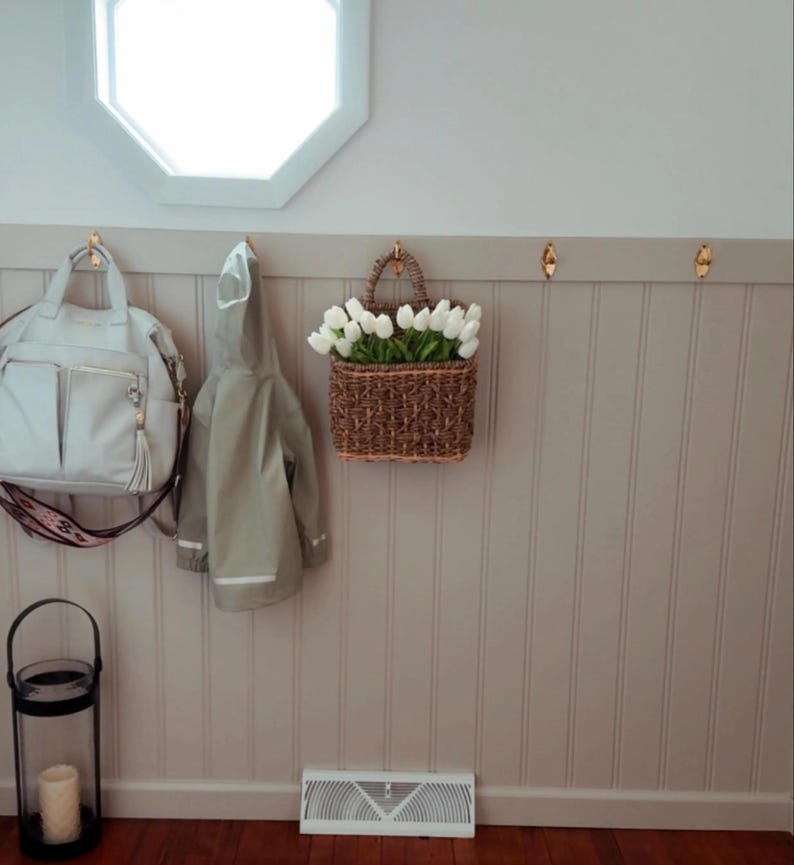 May include: A light gray handbag with a patterned strap hangs on a brass hook next to a light green raincoat. A woven basket filled with white tulips is suspended from a hook. A black lantern with a candle sits on the floor.