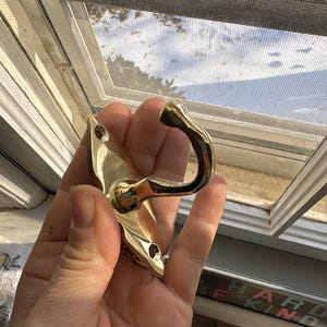 May include: A shiny gold-colored metal hook with a curved design. The hook has a decorative base with screw holes for mounting. The hook is held in a hand near a window.