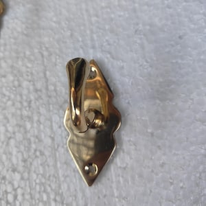 May include: A gold-colored metal hook with a decorative, diamond-shaped backplate. The hook has a curved design and a small hole at the bottom for mounting. The hook is likely for hanging items.