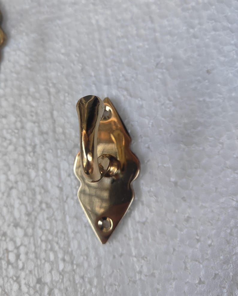 May include: A gold-colored metal hook with a decorative, diamond-shaped backplate. The hook has a curved design and a small hole at the bottom for mounting. The hook is likely for hanging items.