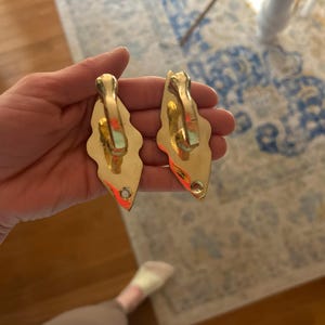 May include: Two ornate, gold-colored metal hooks. Each hook has a decorative, leaf-like shape with a curved top and a small hole at the bottom. The hooks are likely for hanging items and have a vintage or antique aesthetic.