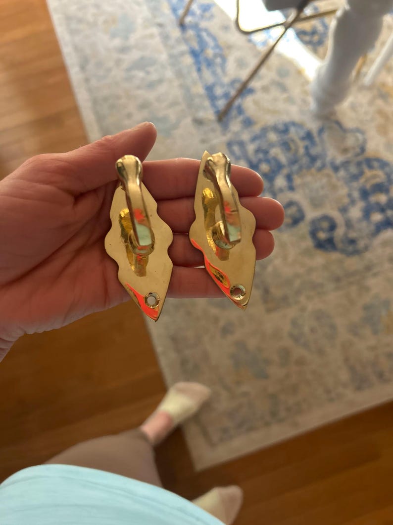 May include: Two ornate, gold-colored metal hooks. Each hook has a decorative, leaf-like shape with a curved top and a small hole at the bottom. The hooks are likely for hanging items and have a vintage or antique aesthetic.
