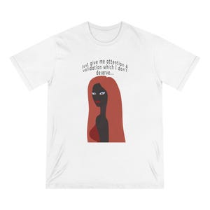 May include: White t-shirt featuring a graphic of a woman with long red hair and the text "Just give me attention & validation which I don't deserve...". The woman's skin is dark, and she is wearing a red top.