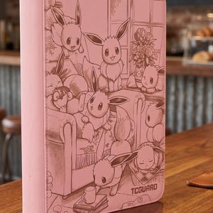 Pode incluir: Um fichário de cartas colecionáveis rosa claro com uma ilustração detalhada de vários personagens Eevee em um ambiente aconchegante. A capa exibe várias figuras de Eevee em móveis e no chão, com a marca TCGUARD impressa na parte inferior.