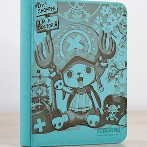 Puede incluir: Una carpeta para tarjetas coleccionables de color turquesa con un gráfico del personaje Dr. Chopper de One Piece. La cubierta muestra el texto Dr. CHOPPER e I'M A DOCTOR! junto a un emblema de calavera, flores de cerezo y el logotipo TCGUARD.