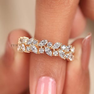 May include: A gold ring with a leaf design, set with multiple clear gemstones. The ring is held up by a person's finger, showcasing the intricate details of the jewelry. The ring's design is elegant and eye-catching.