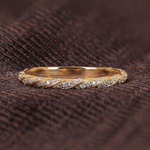 Op de afbeelding: Een goudkleurige ring met een gedraaid ontwerp, bezet met kleine, sprankelende diamanten. De ring is geplaatst tegen een donkerbruine, getextureerde achtergrond, die de schittering benadrukt. De band is ongeveer 2 mm breed.