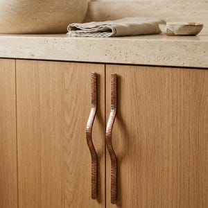 May include: Close-up of a bathroom vanity with two copper-colored, wavy cabinet pulls. The cabinet doors are made of light-colored wood. The pulls have a textured surface. The bathroom sink and faucet are visible in the background.