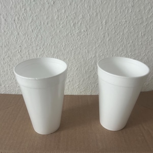 May include: Two white, tapered foam cups are displayed on a brown surface. The cups have a smooth, clean appearance and are empty. The background is a textured white wall.