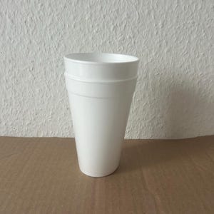 Könnte beinhalten: Ein weißer, konisch zulaufender Schaumstoffbecher mit zwei horizontalen Rillen am oberen Rand. Der Becher ist leer und steht auf einer braunen Oberfläche. Der Hintergrund ist eine strukturierte weiße Wand.