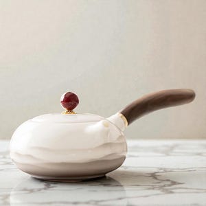 Puede incluir: Una tetera de cerámica blanca con un asa de madera marrón oscuro y una pequeña tapa redonda roja. La tetera tiene una forma redondeada con una banda marrón claro alrededor de la base. El asa está sujeta con una banda dorada.