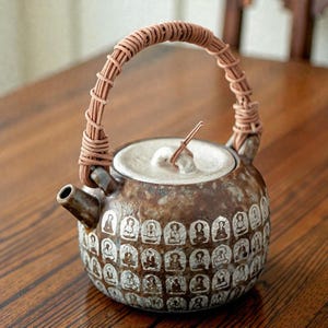 May include: A brown ceramic teapot with a woven handle and a lid. The body of the teapot is decorated with a repeating pattern of white figures. The spout is on the left side of the teapot.
