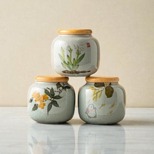 Puede incluir: Tres tarros de cerámica con tapas de madera están apilados. Cada tarro es de color verde pálido con un esmalte craquelado y presenta un diseño pintado a mano diferente: flores, un conejito y ramas florales. Los tarros son decorativos.