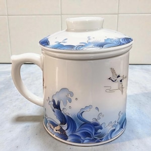 Könnte beinhalten: Weißer Keramikbecher mit passendem Deckel, verziert mit blauen Wellen- und Kranichmotiven. Der Becher hat einen Henkel und ist für Tee oder Kaffee konzipiert. Der Deckel hat einen kleinen Knopf. Der Becher ist etwa 10 cm hoch.
