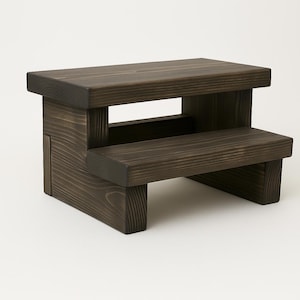 May include: A dark brown wooden step stool with two steps. The step stool is made of solid wood and has a simple, sturdy design. The top step is larger than the bottom step. The wood grain is visible.