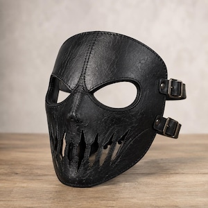 May include: A black textured mask featuring a wide eye opening and a jagged, tooth-like cutout design across the mouth area. The mask is constructed with visible stitching along the center and edges, and includes two adjustable side straps with metal buckles.