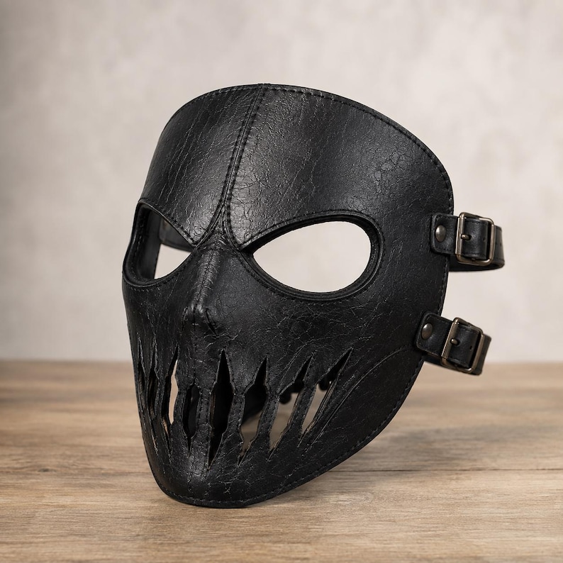 May include: A black textured mask featuring a wide eye opening and a jagged, tooth-like cutout design across the mouth area. The mask is constructed with visible stitching along the center and edges, and includes two adjustable side straps with metal buckles.