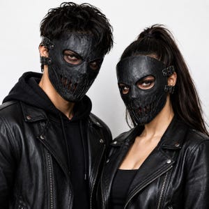 May include: Two black textured leather masks featuring eye cutouts and vertical slit detailing over the mouth area. Each mask is secured with adjustable side straps and metal buckles, covering the upper face and nose while leaving the eyes visible.