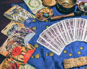 May include: A collection of tarot cards, a singing bowl, a candle, and other spiritual items arranged on a blue cloth. The cards feature colorful illustrations and are fanned out. A bundle of sage is also present.