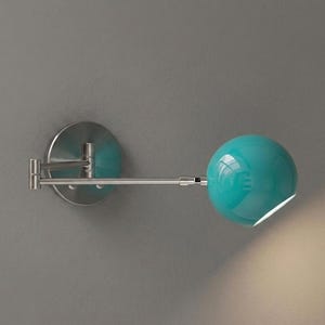 May include: A modern wall-mounted lamp with a teal spherical shade. The lamp features a brushed metal finish and an adjustable arm. The round base is also brushed metal. The lamp is designed to provide focused lighting.