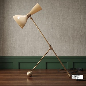 May include: A table lamp with a dual-cone cream shade and a slender brass-toned metal frame. The lamp features an adjustable arm with a central pivot point and a spherical base weight. The text ORREN GLOW is visible on a small white label.