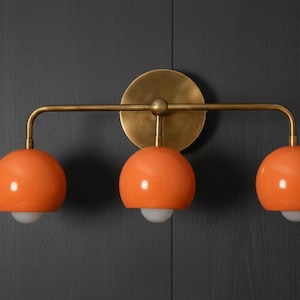 Può includere: Un apparecchio per il trucco a tre luci con paralumi a globo arancioni e finitura in ottone. L'apparecchio è montato su una parete grigio scuro. Ogni globo ha una lampadina bianca.