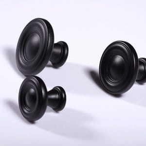 Op de afbeelding: Drie zwarte kastknoppen met een cirkelvormig ontwerp. De knoppen zijn gemaakt van een solide materiaal en hebben een matte afwerking. De knoppen zijn gerangschikt op een wit oppervlak, met schaduwen die diepte toevoegen.