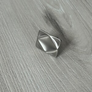 May include: A rectangular cabinet knob with a brushed silver finish, featuring a geometric design with four triangular facets that meet at a central point, creating a faceted, three-dimensional surface with clean, sharp edges and a metallic sheen.