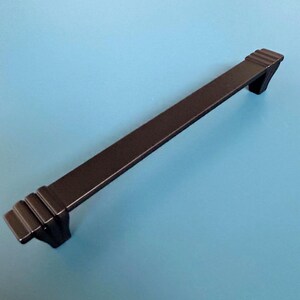 Könnte beinhalten: Ein schwarzer Schrankgriff mit einem rechteckigen Stab und dekorativen Enden. Die Hardware befindet sich vor einem hellblauen Hintergrund. Die Enden des Griffs haben ein abgestuftes Design.