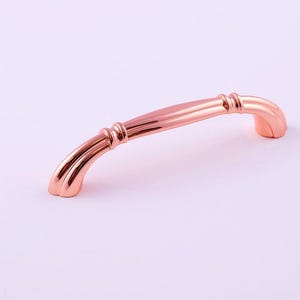Può includere: Maniglia curva per mobili in oro rosa. La maniglia ha una finitura lucida ed è posta su uno sfondo bianco. L'hardware è probabilmente per un cassetto o un'anta dell'armadio. La maniglia è lunga circa 15 cm.
