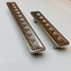 May include: Two brushed nickel cabinet pulls with a rectangular shape and a textured, geometric pattern. One pull is longer than the other. The pulls are set against a white background.