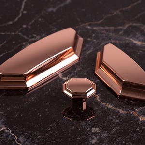 Può includere: Un set di tre elementi di ferramenta per mobili in oro rosa. Il set comprende due maniglie allungate e un singolo pomello ottagonale. La ferramenta ha una finitura metallica riflettente ed è esposta su una superficie scura e marmorizzata.