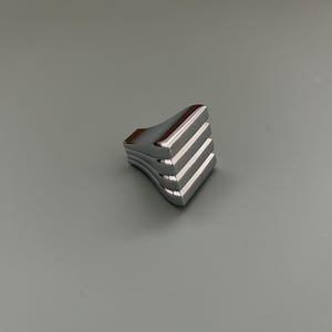 Peut inclure: Un petit bouton de meuble chromé avec un design superposé unique. Le bouton a un dessus incurvé et une série de lignes horizontales, créant une esthétique géométrique moderne. La surface polie reflète la lumière, ajoutant une touche d'élégance.