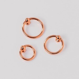 Peut inclure: Trois boucles d'oreilles créoles en or rose avec un petit accent en forme de boule. Les boucles d'oreilles sont disposées sur une surface blanche, mettant en valeur leur design circulaire et leur finition métallique.