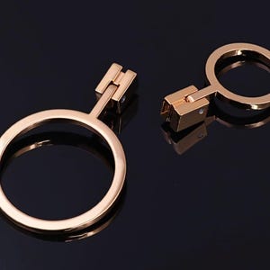 Può includere: Due chiusure metalliche color oro a forma di anello, con una cerniera rettangolare e un piccolo accento quadrato. Le chiusure sono esposte su una superficie scura e riflettente, che mostra la loro finitura lucida e il design.